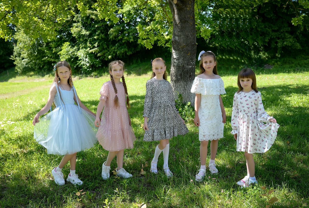 Five young girls in colorful summer dresses standing on grass under a tree.